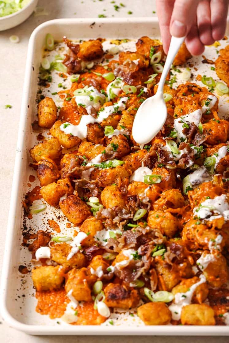 Loaded tater tots being garnished with sour cream on a sheet pan.