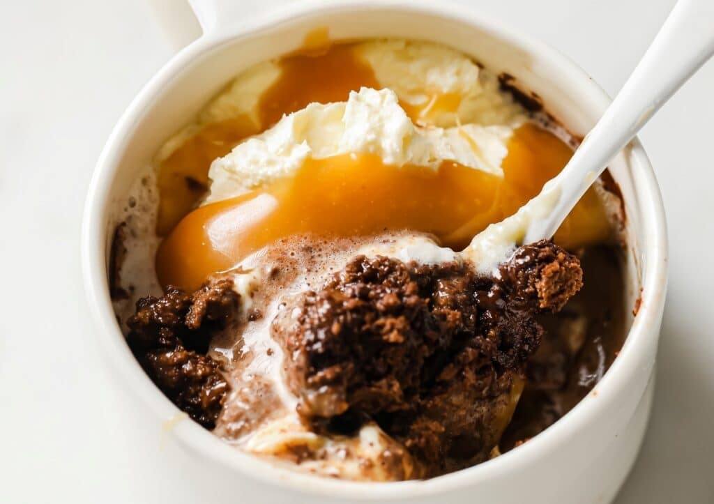 A ceramic mug filled with a layered dessert, featuring chocolate cake, ice cream, and caramel sauce, is shown with a spoon dipped inside.