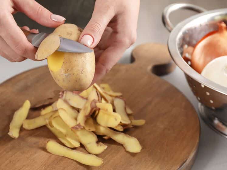 Woman peeling fresh potato with knife at light table indoors, closeup