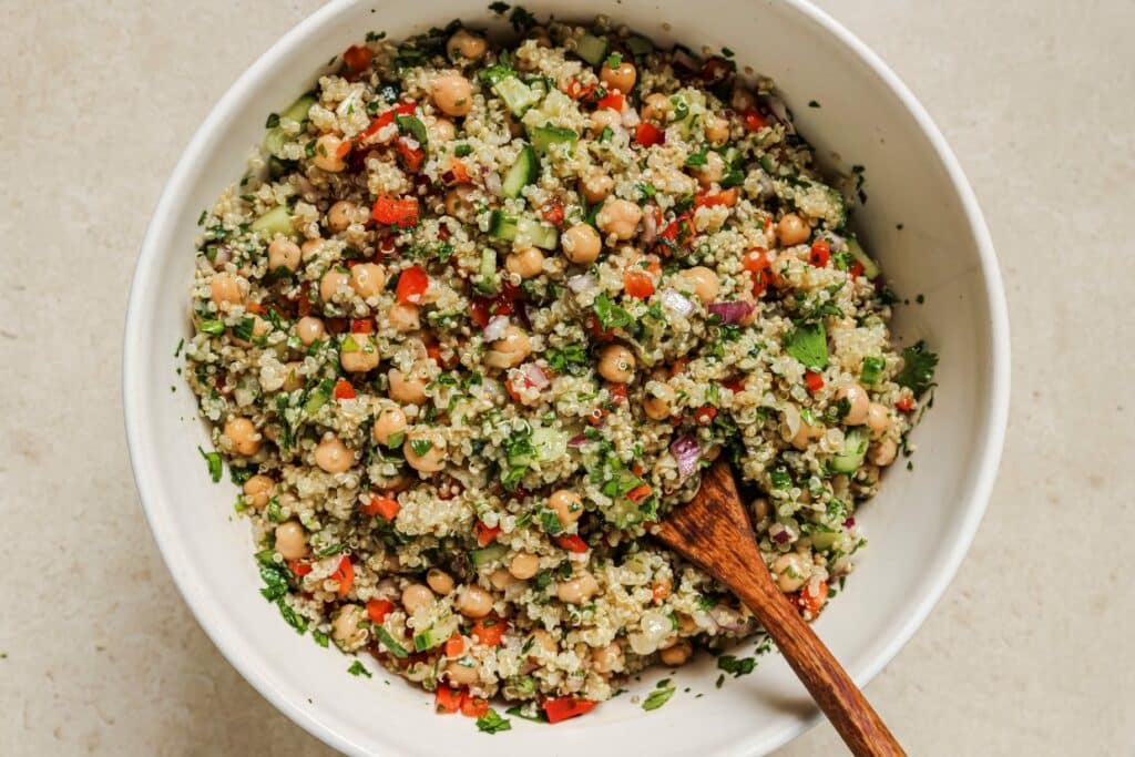 A bowl of quinoa salad with chickpeas, chopped vegetables, herbs, and a wooden spoon.