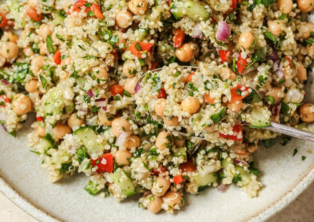 A close-up of a quinoa salad with chickpeas, chopped vegetables, and fresh herbs, being scooped with a spoon.