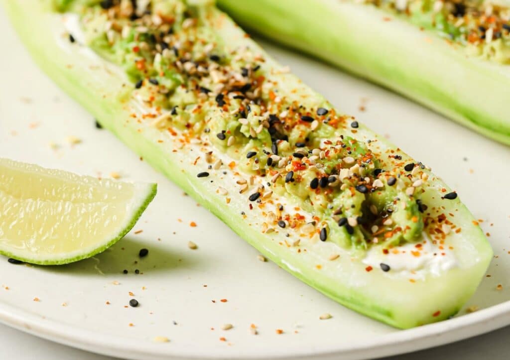 Cucumber boats filled with creamy topping, garnished with everything bagel seasoning, and served with a lime wedge on a white plate.