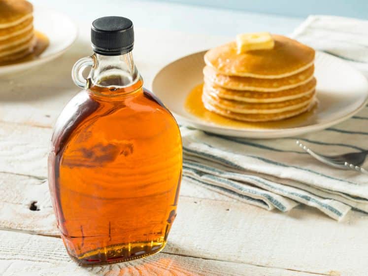 Glass jar of maple syrup in front of a plate of pancakes.