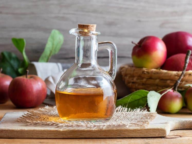 Vinegar in a glass bottle on a cutting board in front of apples.