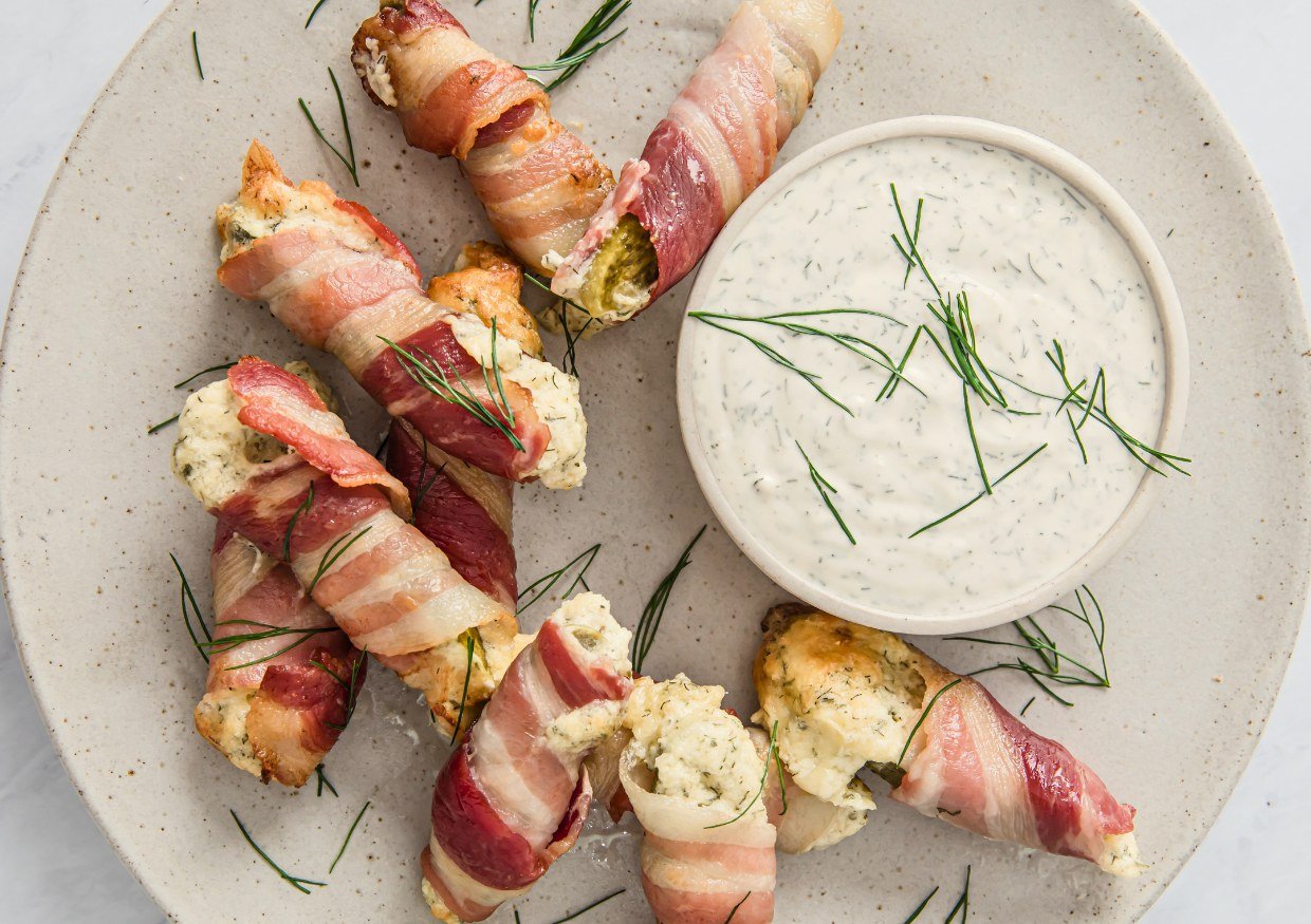 Bacon pickle boats stuffed with cream cheese on a plate next to a bowl of dip.