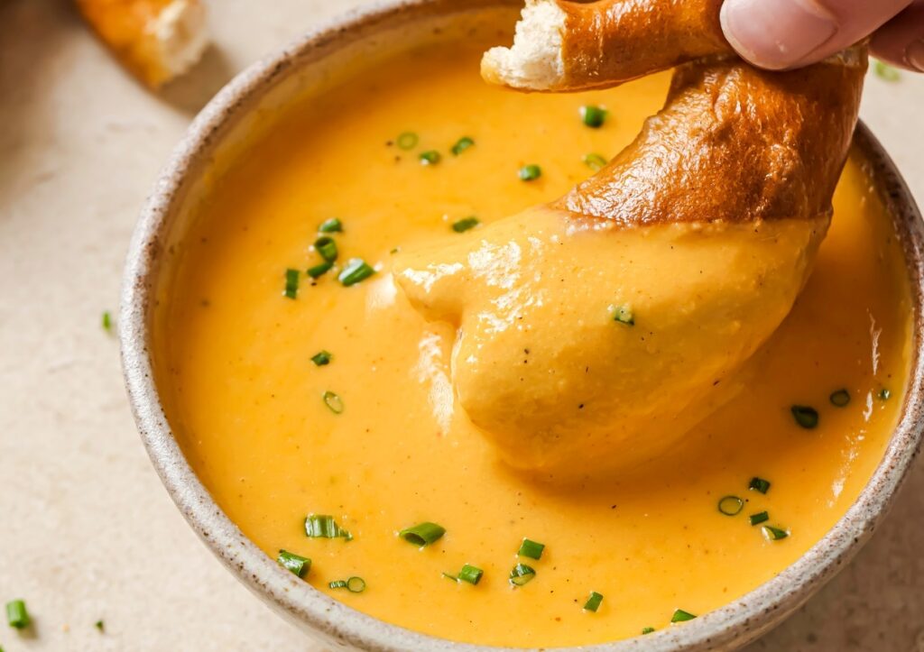 A hand dips a piece of bread into a bowl of creamy orange-yellow soup or sauce, garnished with chopped green herbs.