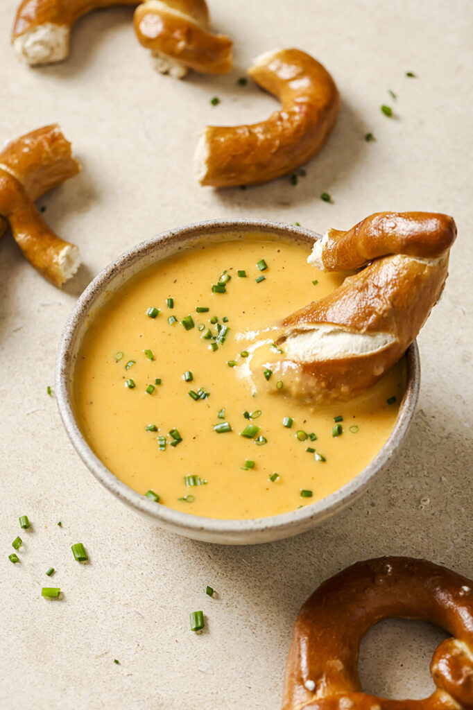 A bowl of beer cheese dip with soft pretzel and soft pretzel pieces all around.