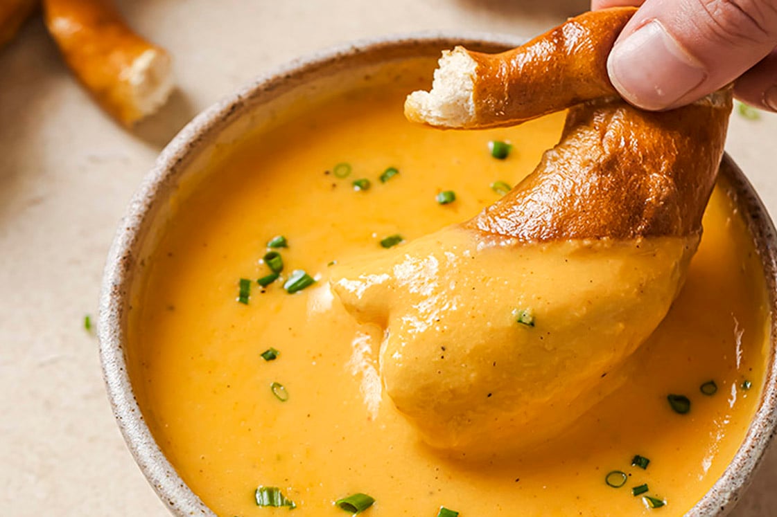 A hand dipping soft pretzel in a bowl of beer cheese dip.