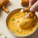 A hand dipping soft pretzel in a bowl of beer cheese dip.