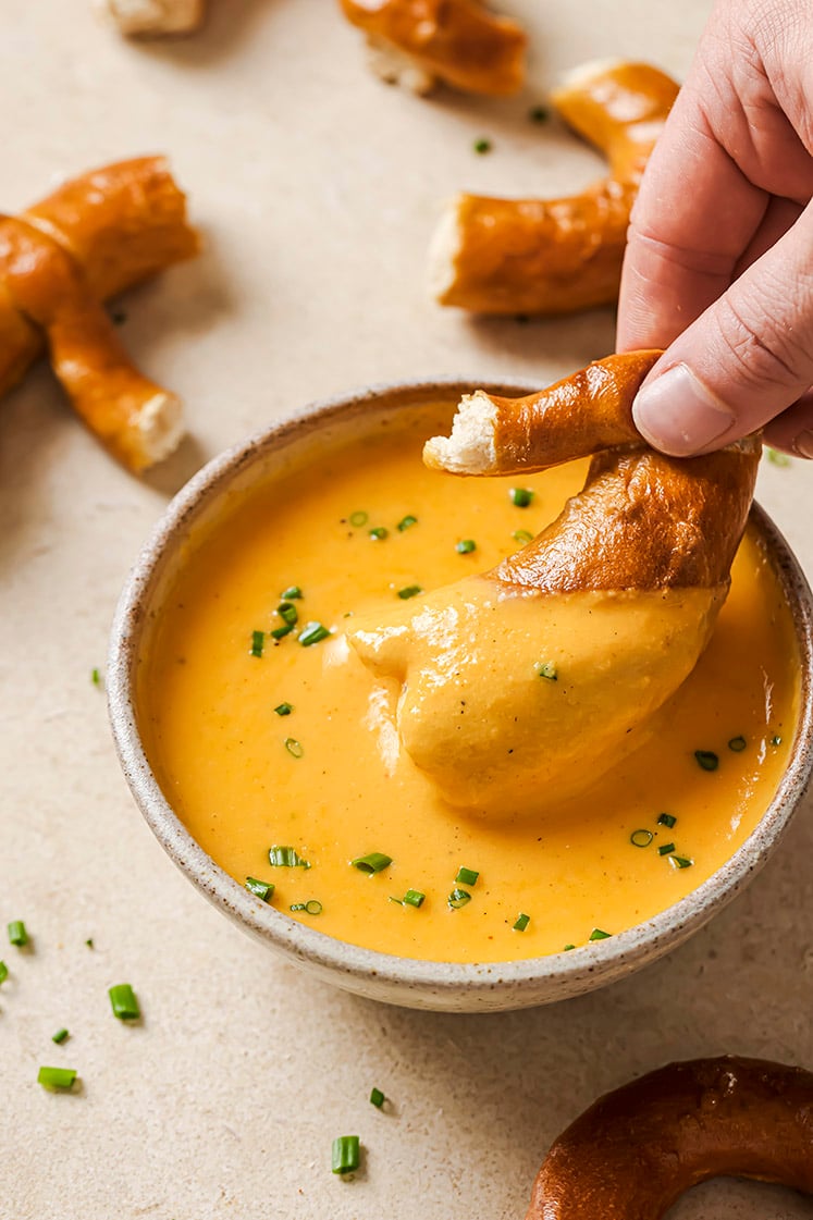 A hand dipping soft pretzel in a bowl of beer cheese dip.