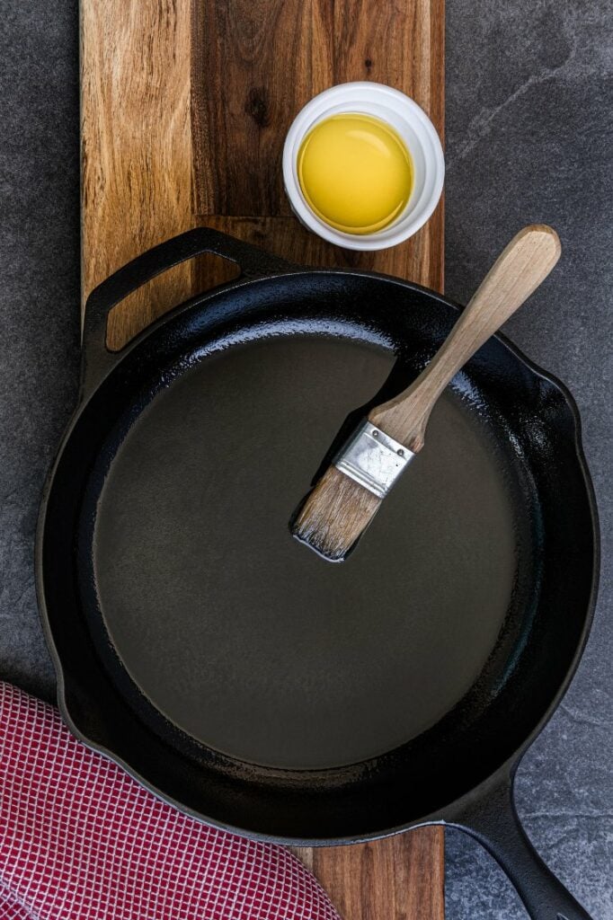 Cast iron skillet with a thin layer of oil being applied by a brush.