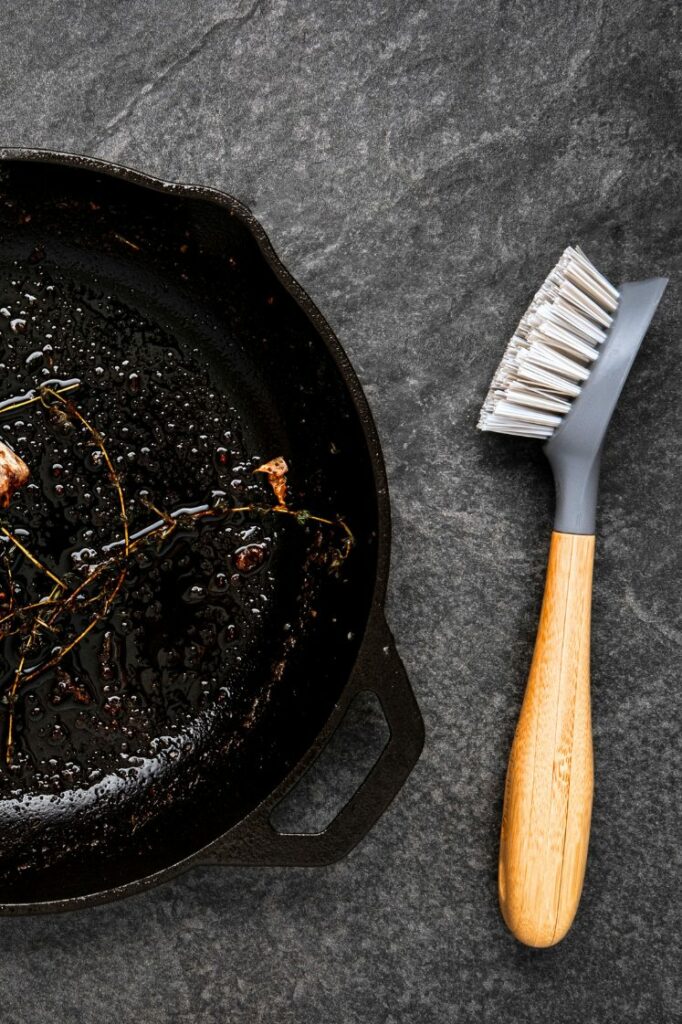 Cast iron skillet with food residue beside a dish brush.