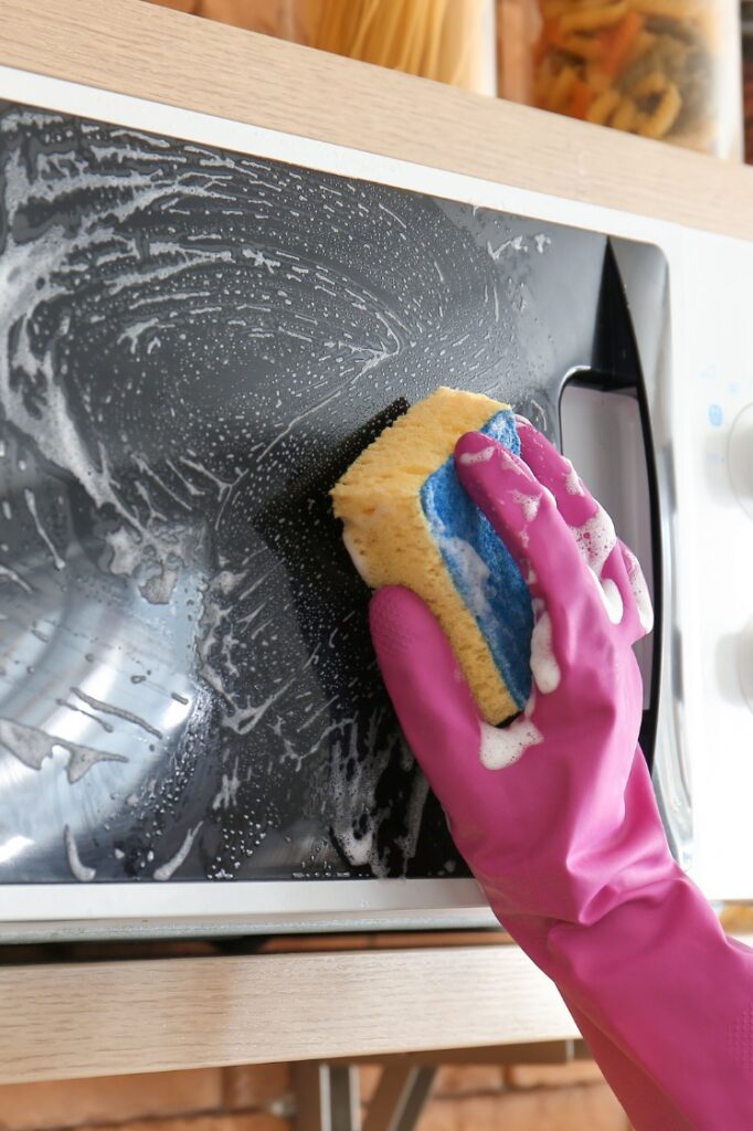 A person cleaning the outside of a microwave with a soapy sponge.