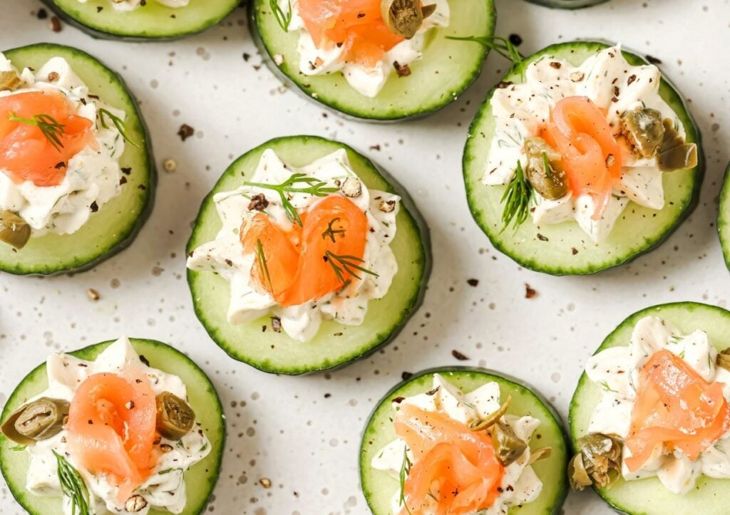 Slices of cucumber topped with a dollop of cream cheese, smoked salmon, capers, and dill, arranged on a white speckled surface.