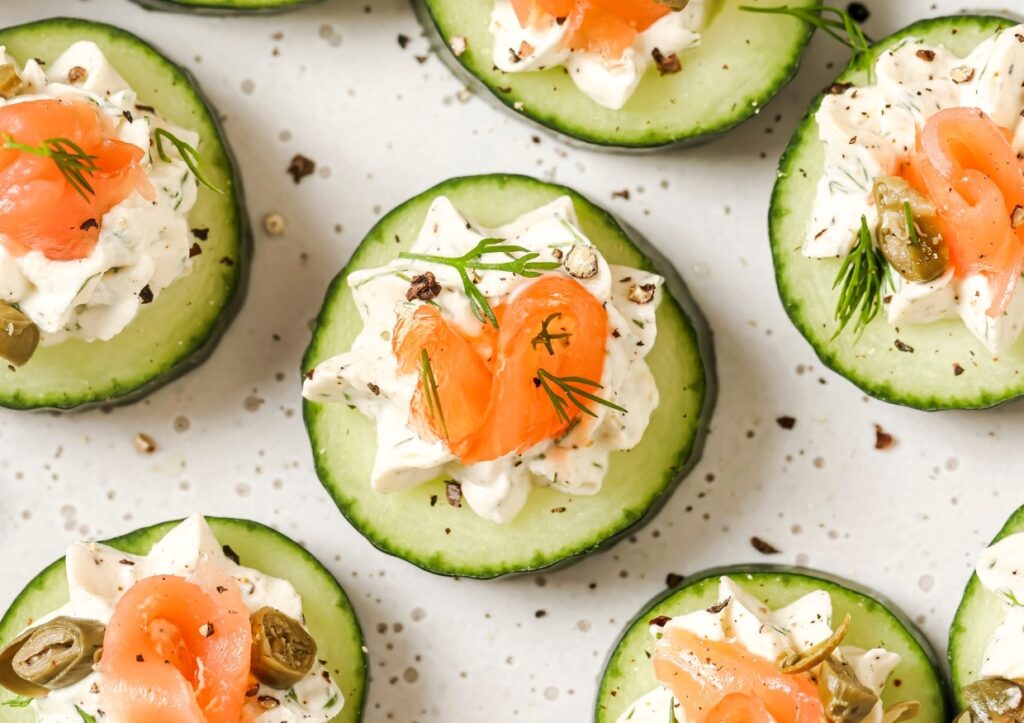 Cucumber slices topped with cream cheese, smoked salmon, dill, capers, and seasoning, arranged on a white surface.