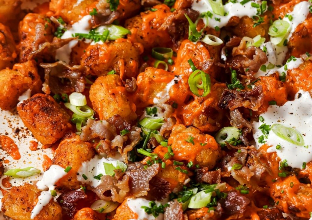 Close-up of loaded tater tots topped with melted cheese, crumbled bacon, sliced green onions, sour cream, and garnished with chopped herbs.