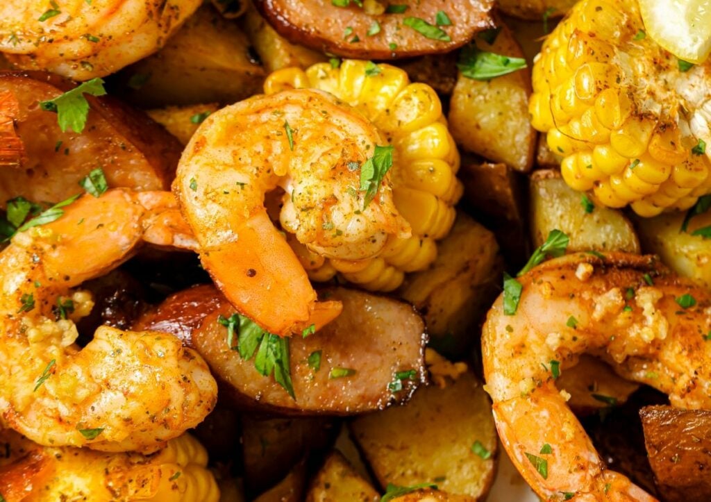 Close-up of a shrimp boil featuring cooked shrimp, corn on the cob, sausage, and potatoes, garnished with herbs.