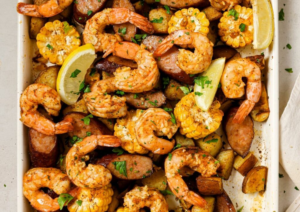 A tray of baked shrimp, sausage, corn on the cob slices, and potatoes garnished with lemon wedges and chopped parsley.