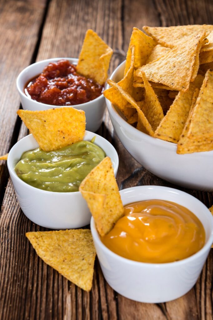 Small bowls with salsa, guacamole, and cheese dip beside a bowl of tortilla chips.