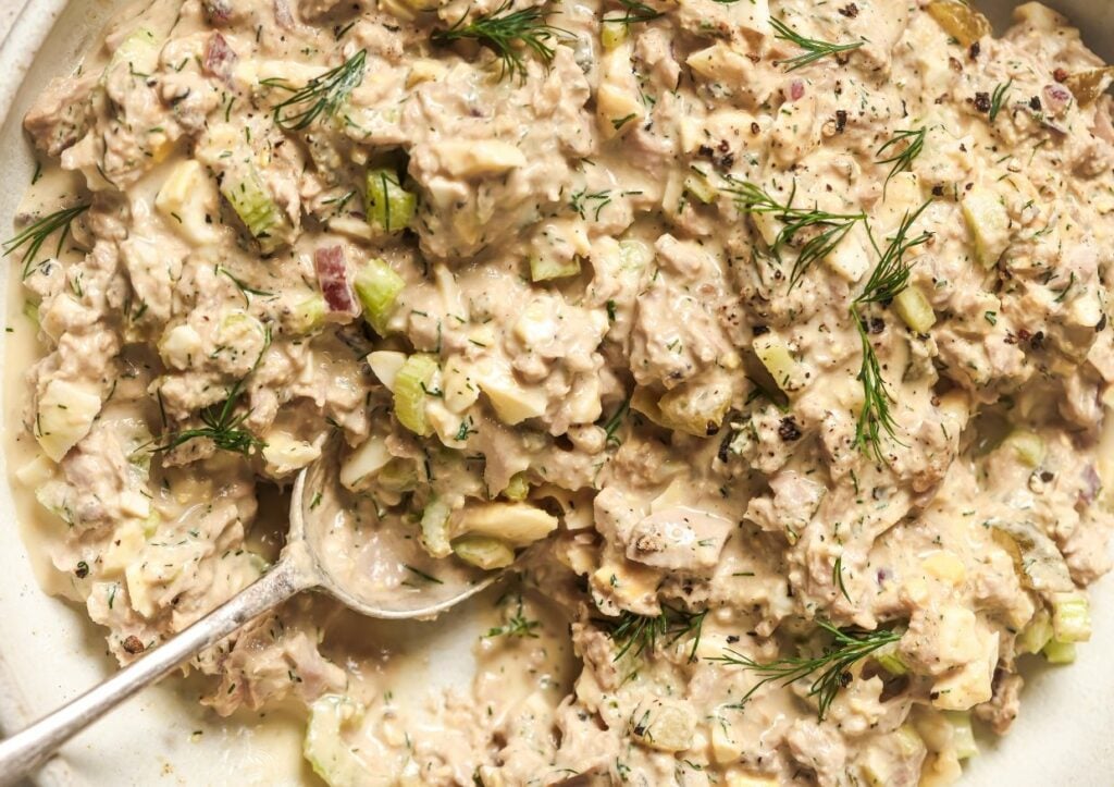 A close-up of a creamy tuna salad with diced celery and garnished with fresh dill, shown with a serving spoon.