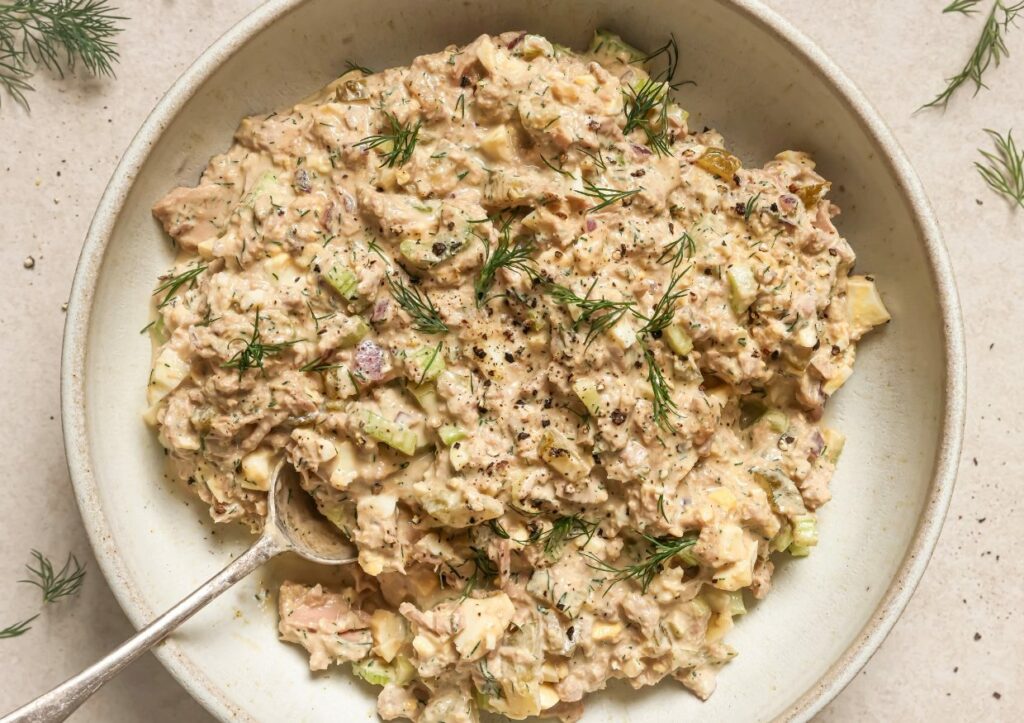 A bowl of tuna salad mixed with chopped celery, dill, and mayonnaise, garnished with fresh dill sprigs, with a spoon resting on the side.