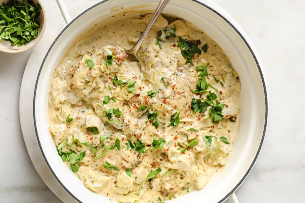 Creamy skinless chicken thighs with artichoke hearts in a skillet with a serving spoon.