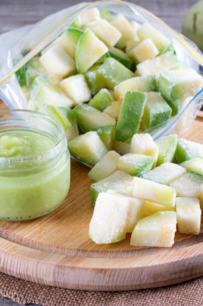 Frozen zucchini cubes and a small jar of zucchini puree on a round cutting board.