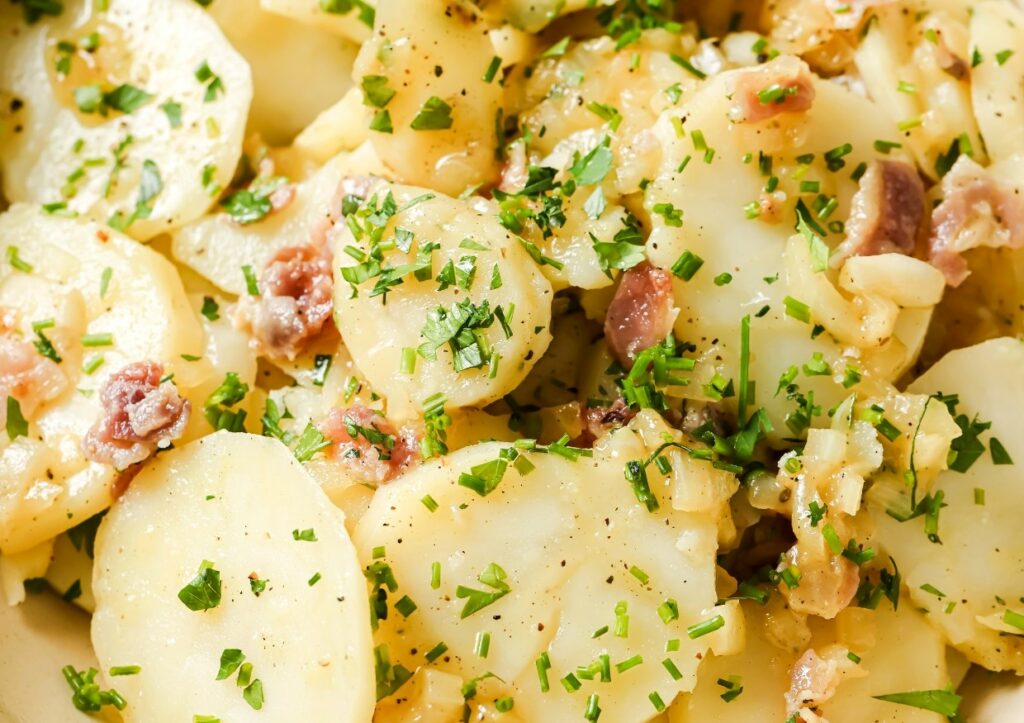 A close-up of a potato salad with chopped herbs, bacon bits, and seasonings.