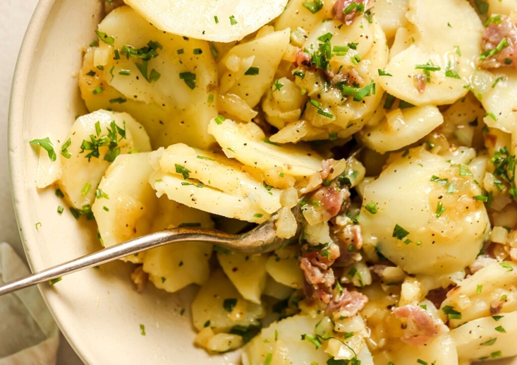 A close-up of a potato salad with slices of potato, chopped parsley, and chunks of meat, with a fork resting in the dish.