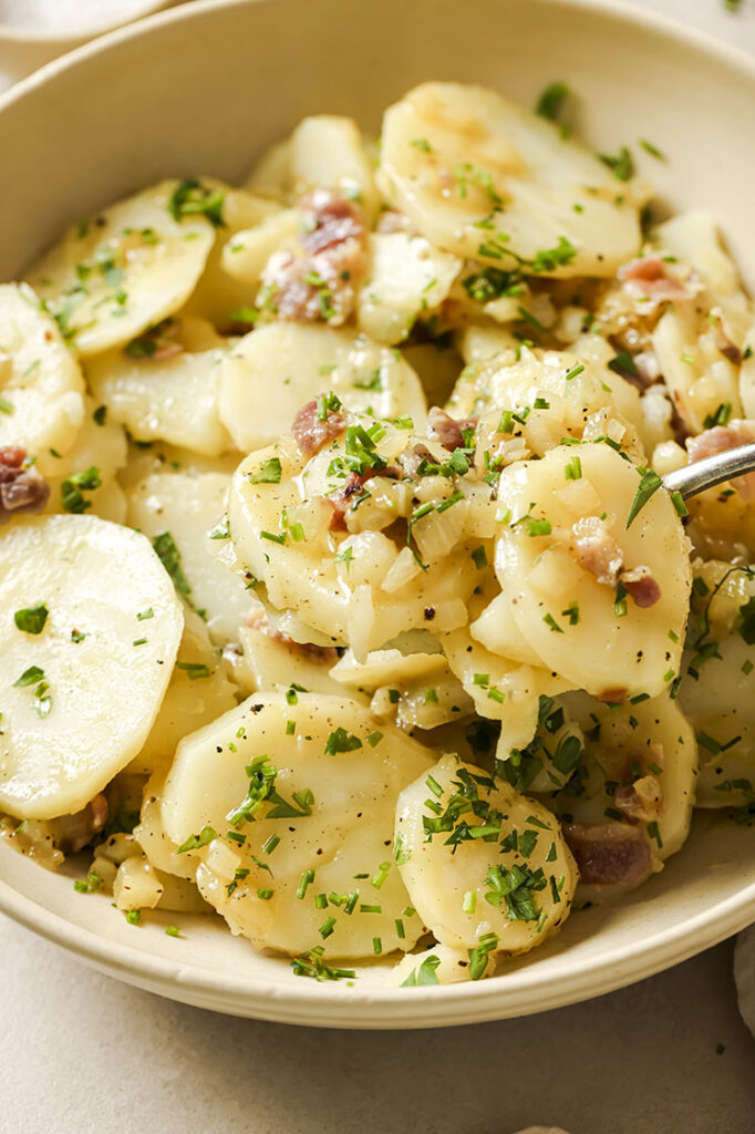 Garnished German potato salad being scooped by a spoon in a bowl.