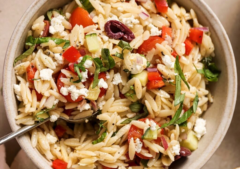 A bowl of orzo pasta salad with chopped tomatoes, olives, cucumbers, red onions, basil, and crumbled feta cheese.