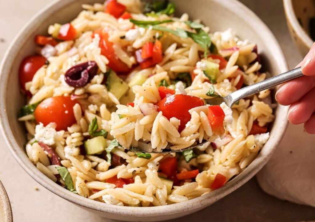 A bowl of orzo pasta salad with cherry tomatoes, cucumbers, red onions, olives, feta cheese, and fresh basil leaves. A spoon is lifting a portion of the salad.