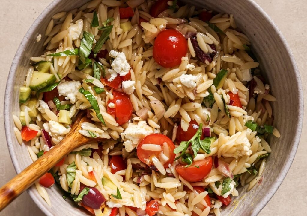 A bowl of orzo salad with cherry tomatoes, cucumbers, red onion, feta cheese, and fresh herbs. A wooden spoon is placed in the bowl.