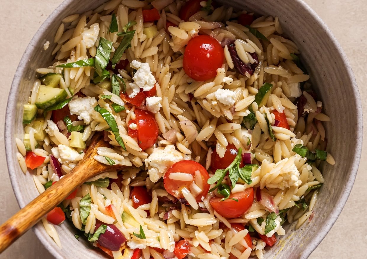 A bowl of orzo pasta salad with tomatoes, olives, cucumbers, red onion, basil, and feta.