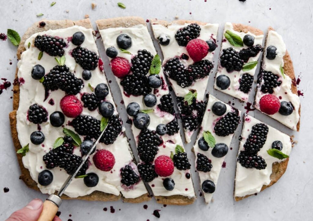 A fruit pizza being sliced into pieces with a knife.