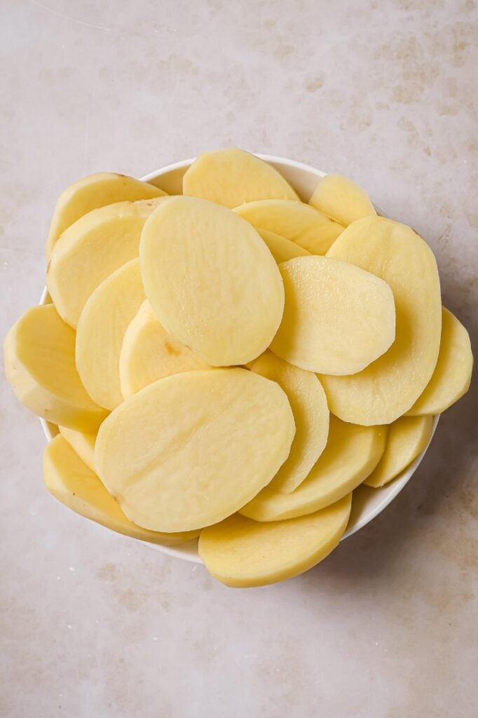 A white bowl filled with thinly sliced raw potatoes.