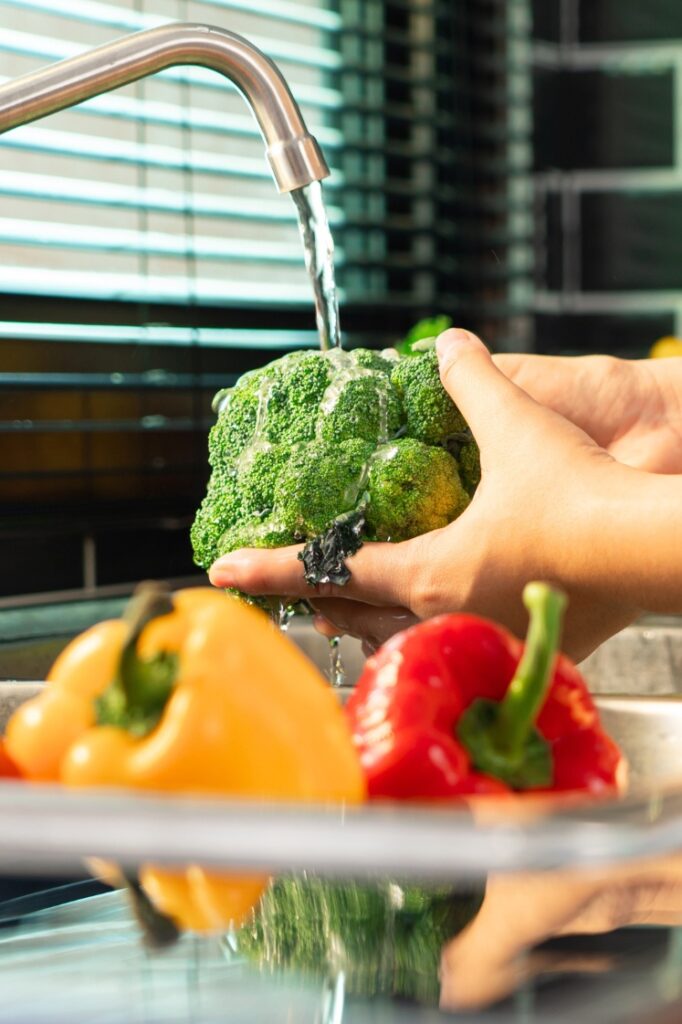 A person washing a head of broccoli and bell peppers.