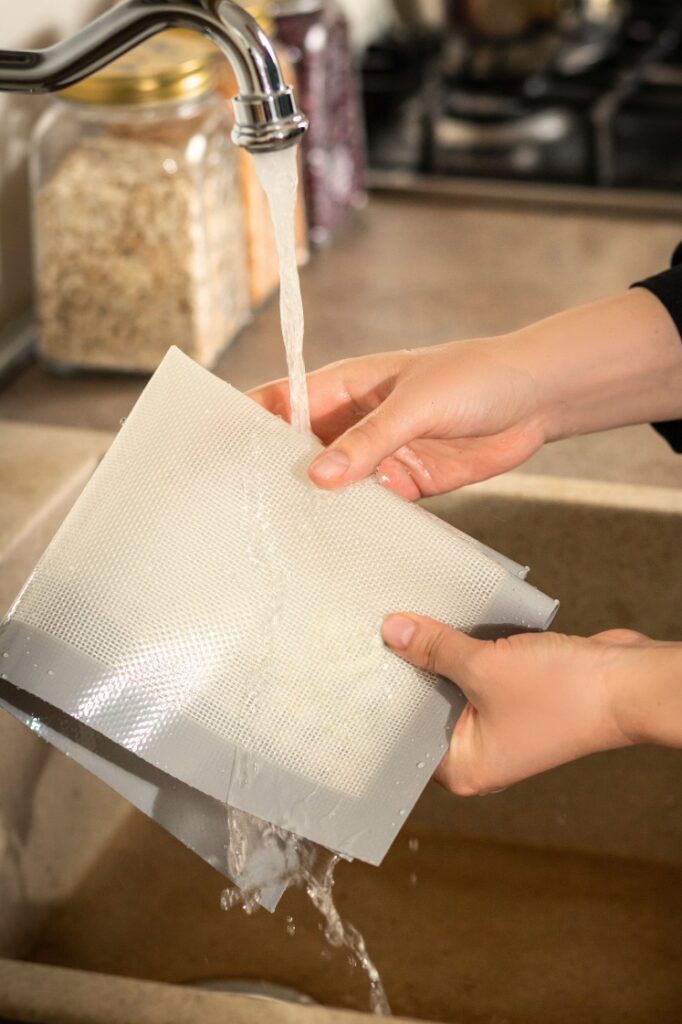 A person washing a silicone mat under a running faucet in a kitchen sink.