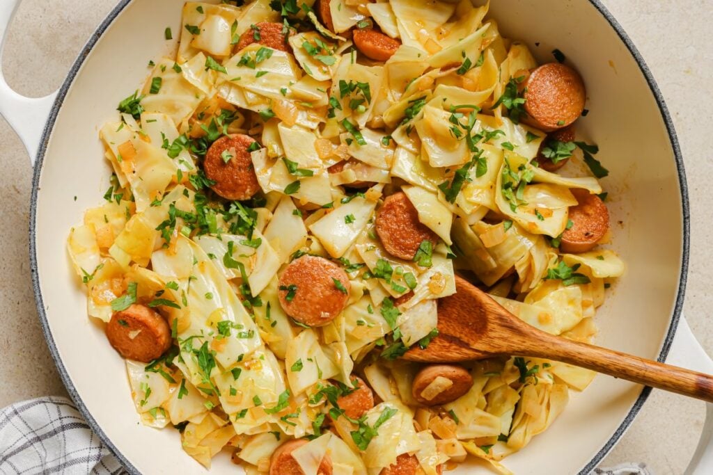 Cabbage and sausage in a skillet with a wooden spoon.