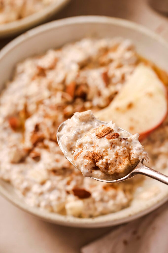 A spoonful of overnight apple pie oats, over the same bowl that’s garnished with chopped almonds, apple slices, cinnamon and maple syrup.