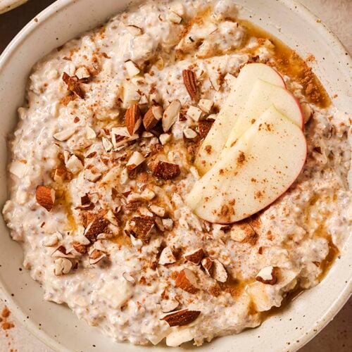 A bowl of overnight apple pie oats garnished with chopped almonds, apple slices, cinnamon and maple syrup.