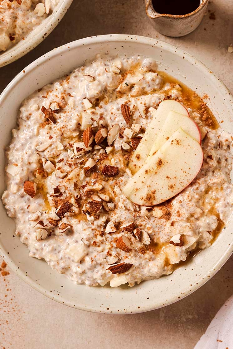 A bowl of overnight apple pie oats garnished with chopped almonds, apple slices, cinnamon and maple syrup.