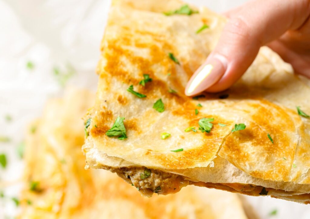 Close-up of a hand holding a triangular piece of quesadilla garnished with green herbs.