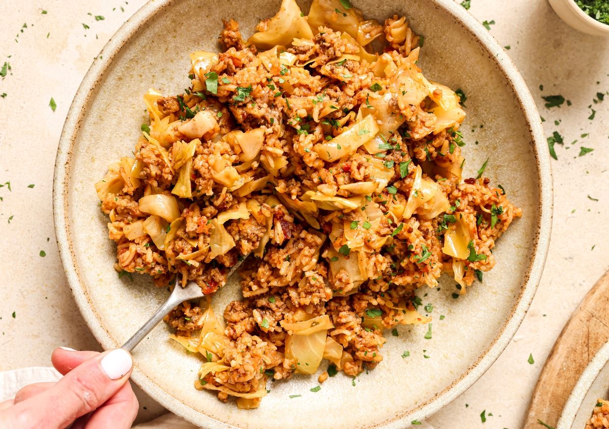 A hand holding a spoon serves a portion of fried rice mixed with beef, cabbage, and garnished with chopped herbs from a beige bowl.