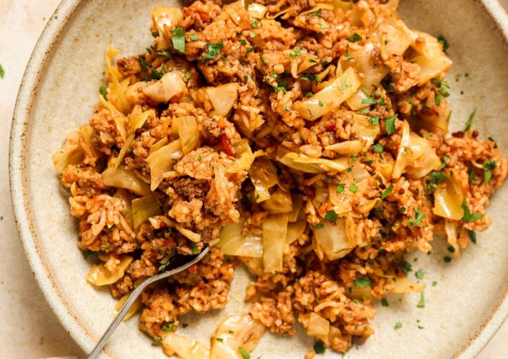 A close-up of a bowl featuring a dish of cooked cabbage, rice, and ground meat, garnished with chopped herbs, with a fork placed on the side.