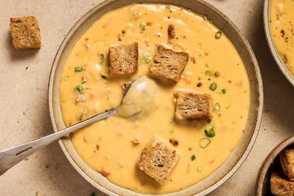 A bowl of creamy soup topped with croutons and chopped green onions, with a spoon in the bowl. Another bowl of soup is partially visible on the right.