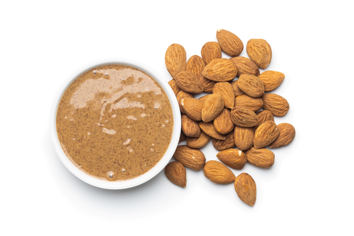 A small bowl of almond butter is placed next to a heap of whole almonds on a white background.