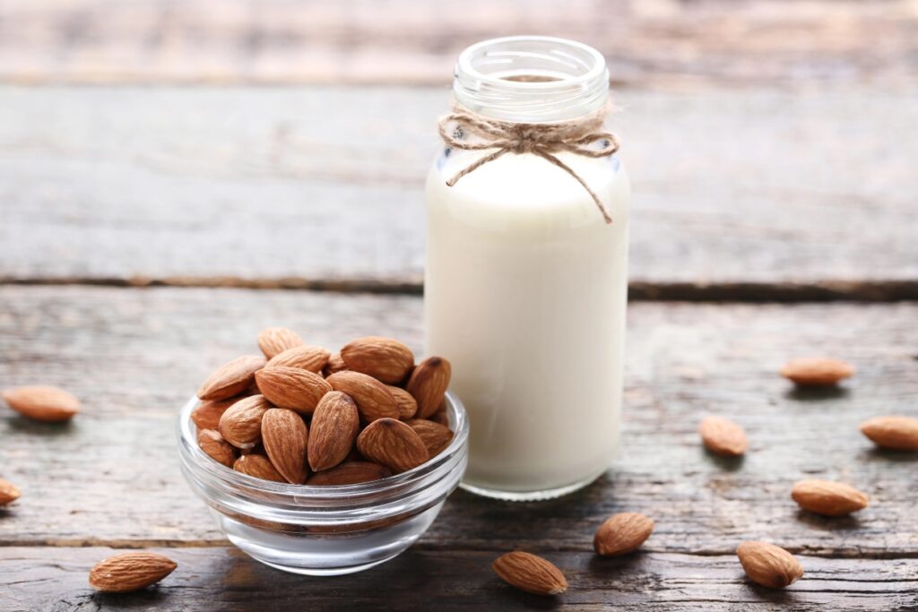 A glass bottle filled with almond milk, tied with a string, stands next to a small glass bowl of whole almonds on a rustic wooden surface. Scattered almonds are visible around the bowl.