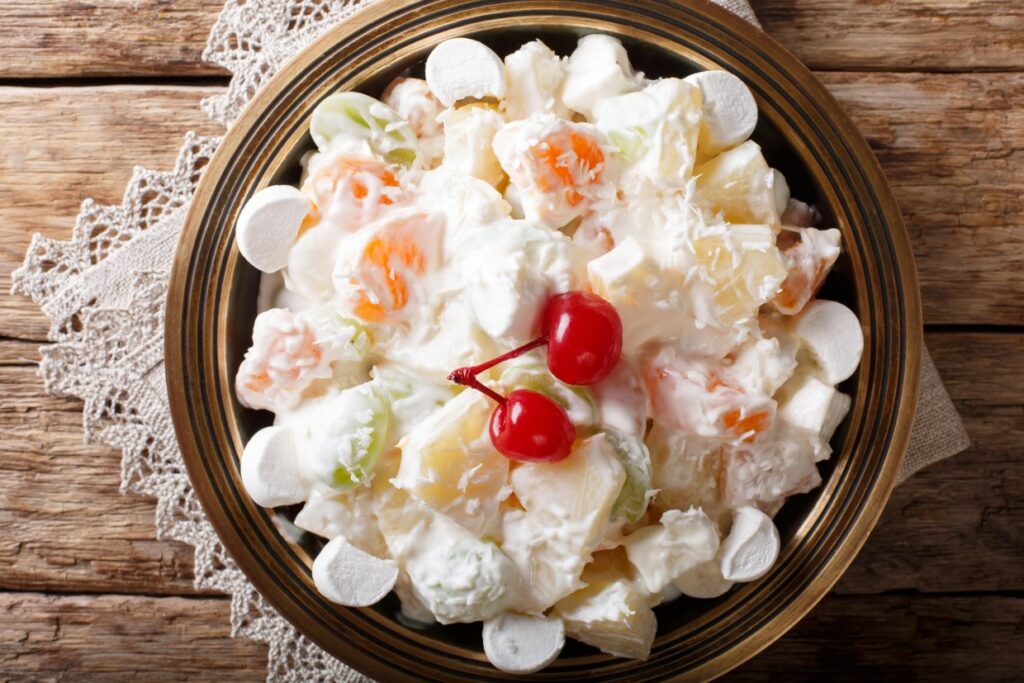 A bowl of creamy ambrosia salad with pineapple chunks, colorful fruit pieces, mini marshmallows, and topped with two maraschino cherries, served on a lace tablecloth over a wooden table.