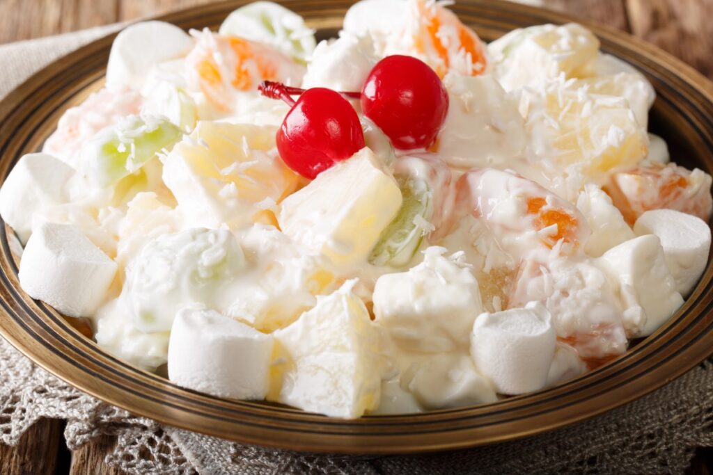 A bowl of ambrosia salad containing diced fruits, marshmallows, and a creamy dressing, garnished with two bright red cherries. The colorful fruit pieces include pineapple and orange segments.